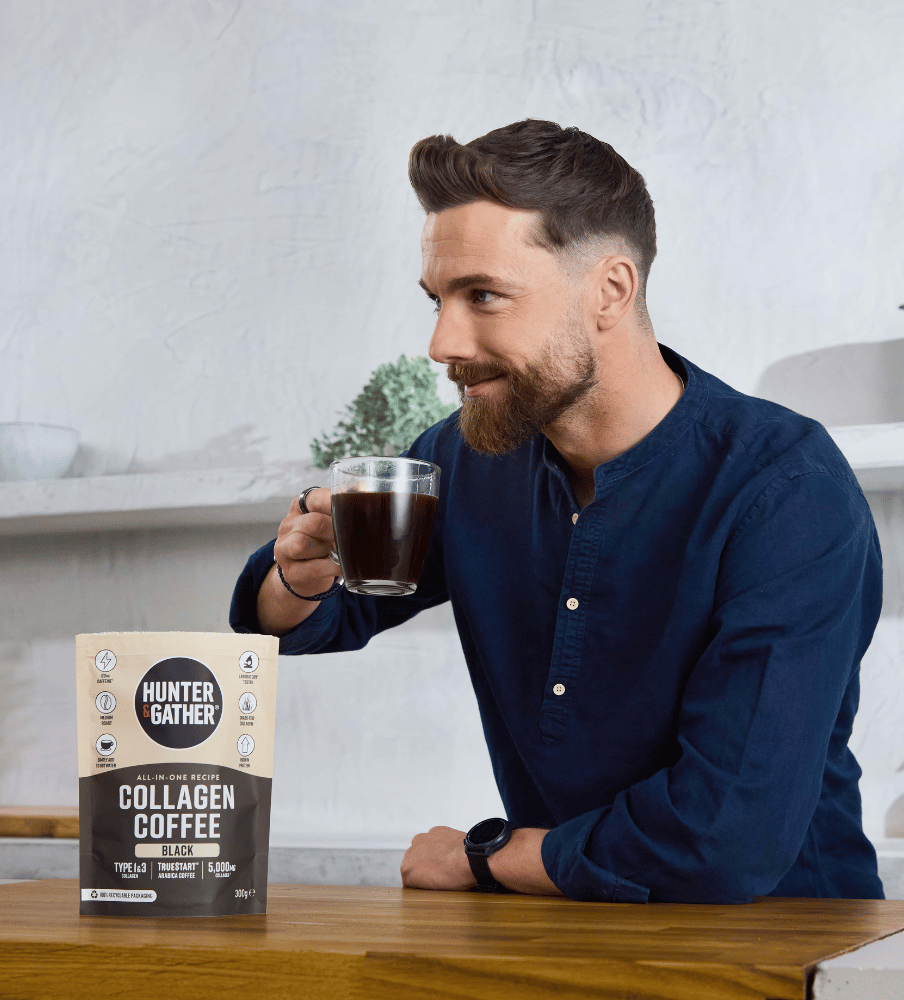 Man holding a glass of coffee next to a container of Hunter Gather collagen coffee on a wooden table.
