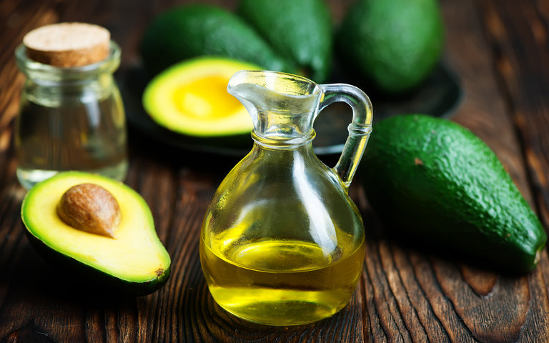 Avocado oil in a decanter surrounded by avocados on a wooden surface