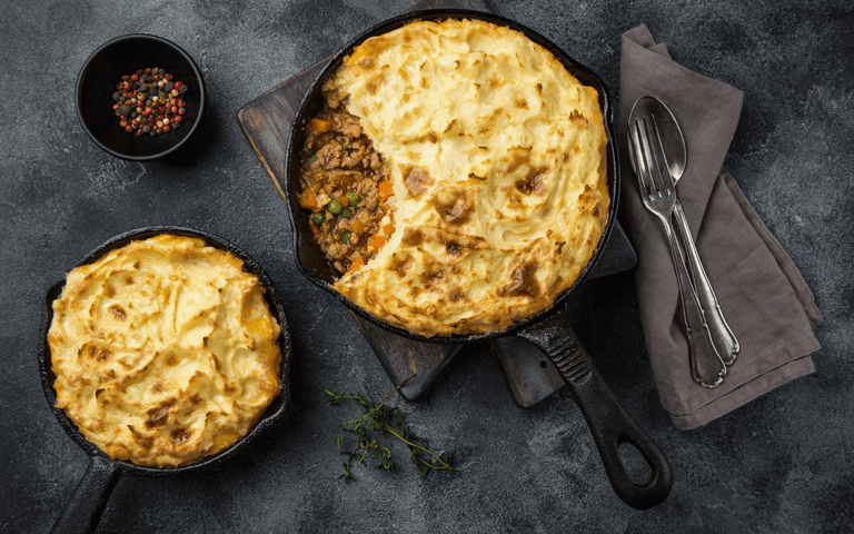 Two shepherd's pies in cast iron pans on a black table with a fork, spoon, and napkin on the side 