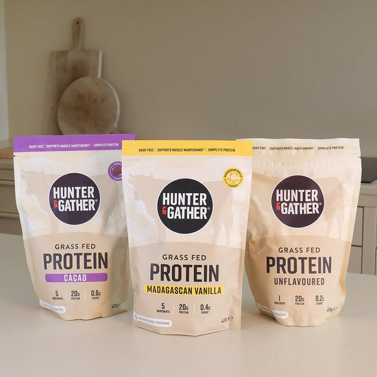 Three stand-up pouches of Hunter Gather Grass Fed Protein powder arranged on a light countertop in a kitchen setting. The pouches are labeled Cacao, Madagascar Vanilla, and Unflavoured, with different accent colors on the packaging.